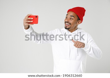 Cheerful young african american man 20s wearing casual streetwear hoodie standing doing selfie shot on mobile phone pointing thumb on himself isolated on white color background. Tattoo translate fun