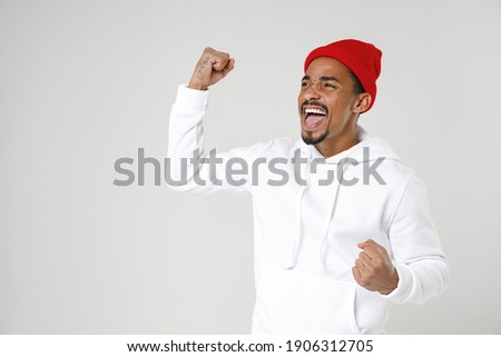 Happy joyful young african man 20s years old in basic streetwear hoodie standing doing winner gesture clenching fists looking aside isolated on white colour wall background studio Tattoo translate fun