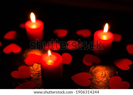 On a black textured background are scattered red hearts and stands three burning red candles, with blurry background, used as a background or texture, soft focus