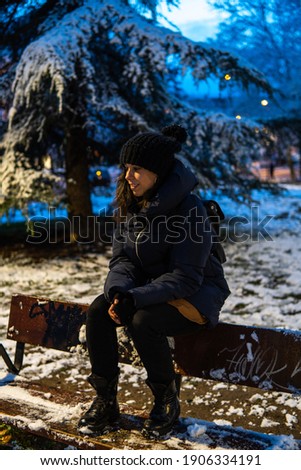 Photo of a young and attractive woman with short hair sitting on a bench surrounded by snow wearing winter clothes and smiling at the camera. Blue sky during the night. Magical landscape