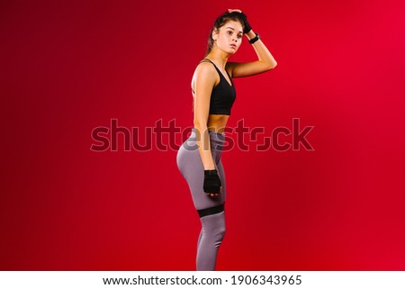 Tired athletic, sweating girl during workout sports exercise with fitness elastic band on her legs. Red background and empty side ad space.