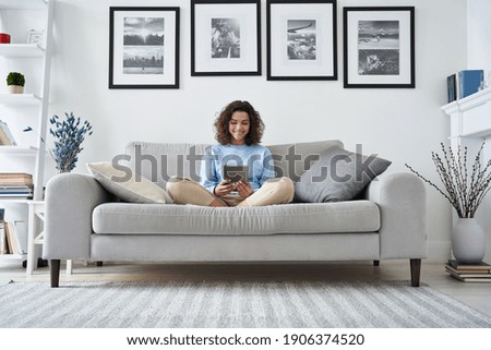 Happy hispanic teen girl holding pad computer gadget using digital tablet technology sitting on couch at home. Smiling young woman using apps, shopping online, reading news, browsing internet on sofa.