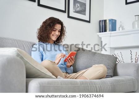 Happy hispanic teen girl holding cell phone using smartphone device at home. Smiling young latin woman blogger subscribing new social media, buying in internet, ordering products online in apps. Royalty-Free Stock Photo #1906374694