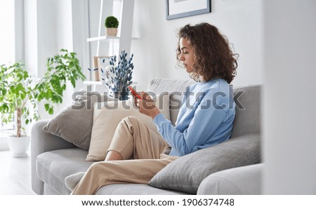 Hispanic teen girl holding cell phone sitting on sofa at home. Young latin woman using smartphone tech mobile apps online services on cellphone device relaxing in apartment living room.