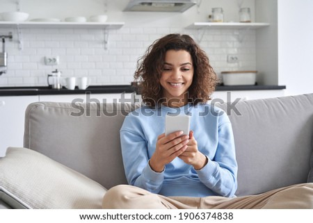 Happy hispanic teen girl looking at smartphone relaxing on couch at home, enjoying using online mobile apps technology, playing games on cell phone, checking messages or social media posts.