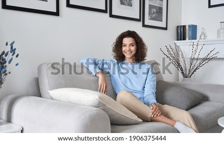 Happy young pretty hispanic woman looking at camera sitting on sofa at cozy home. Happy cheerful casual teen girl, female model posing indoors, relaxing on couch in modern apartment, portrait.
