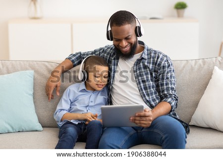 African American Father And Son Using Digital Tablet Wearing Wireless Headphones Watching Cartoons Online Together Sitting On Couch At Home. Family Weekend Leisure, Gadgets And Modern Lifestyle