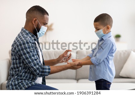 Coronavirus Protection. African Father Spraying Sanitizer On Son's Hands Cleaning Arms At Home, Wearing Protective Face Masks For Covid-19 Prevention. Health Care Concept Royalty-Free Stock Photo #1906391284