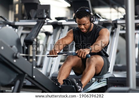 Motivated young black sportsman with wireless headset having workout on rowing machine. Handsome african american man bodybuilder training body on modern block exerciser in gym, copy space Royalty-Free Stock Photo #1906396282