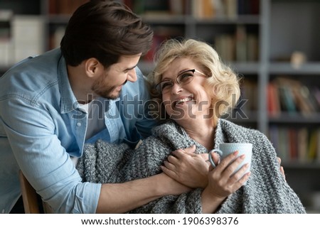 Caring young man covering happy middle aged old mother with plaid, giving cup of hot tea, showing love and devotion at home. Smiling retired woman enjoying peaceful comfort moment with grown son. Royalty-Free Stock Photo #1906398376