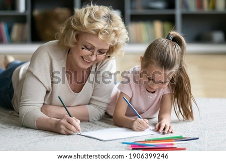 Happy middle aged older woman in eyeglasses lying on floor carpet with smiling adorable preschool little kid girl, enjoying drawing together in paper album, daycare activity, babysitting concept.