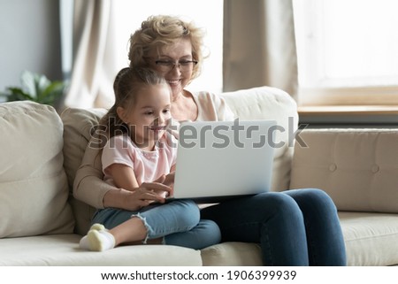 Happy middle aged older grandmother cuddling cute little preschool girl, watching funny cartoons or video on computer, relaxing together on sofa at home, parental control, technology addiction concept