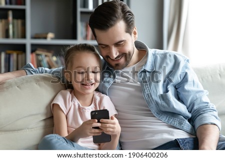 Happy young man cuddling little 6s daughter, playing together on smartphone. Smiling small preschool girl enjoying using mobile applications or watching funny video sitting with caring daddy on sofa. Royalty-Free Stock Photo #1906400206