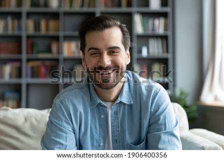 Head shot happy young 30s handsome caucasian man sitting on sofa, looking at camera, involved in web camera conversation with family or friends, enjoying video call distant communication at home. Royalty-Free Stock Photo #1906400356