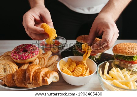 Unhealthy snack, junk food, compulsive overeating. Man overeating unhealthy meals taking hamburger and potato chips from plate Royalty-Free Stock Photo #1906405480