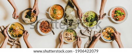 People eating Autumn and Winter creamy vegan soups. Flat-lay of peoples hands with homemade soup and bread slices over white table background, top view. Fall and Winter food menu, vegetarian food Royalty-Free Stock Photo #1906422079