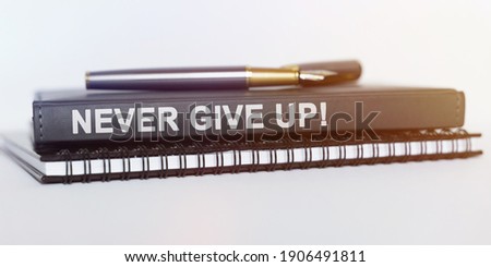 On the table are a notebook, a pen and a book. The book says - NEVER GIVE UP