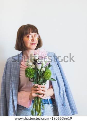 Young woman wearing blue jacket and pink shirt holding beautiful pastel peony bouquet on light gray background