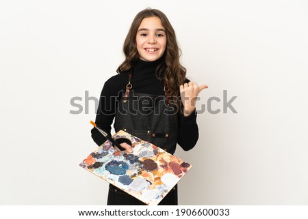 Little artist girl holding a palette isolated on white background pointing to the side to present a product
