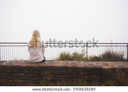 A young blonde Australian female tourist at Govetts Leap Lookout in the Blue Mountains National Park, sits starting into nothing because of whiteout low cloud conditions.