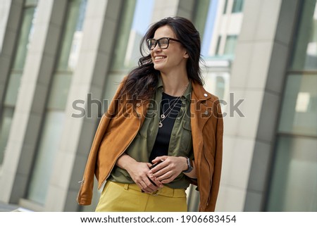 Portrait of a young happy woman holding smartphone, looking aside and smiling while walking on the city street on autumn day, stylish lady standing against blurred urban background