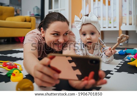 Cute curious little baby girl watching cartoon on mobile phone and playing toys while spending time with happy mother at home