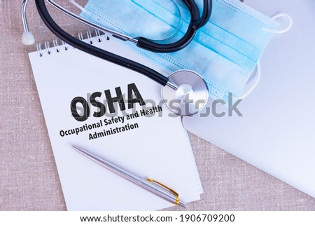 OSHA Occupational Safety and Health Administration, the text is written in a notebook, next to a pen, a disposable medical mask and a laptop on a linen background. Royalty-Free Stock Photo #1906709200