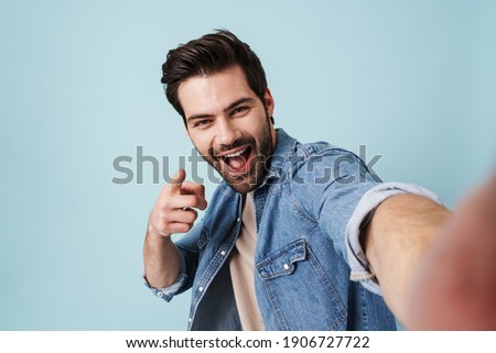 Young excited man pointing finger at camera while taking selfie photo isolated over blue background