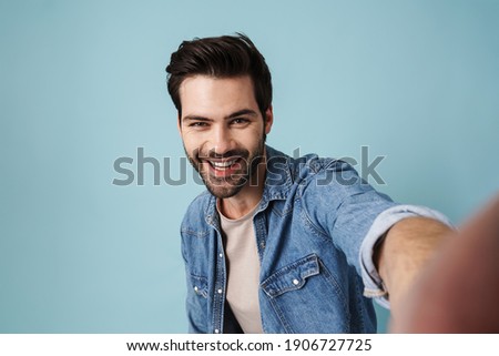 Young handsome happy man smiling while taking selfie photo isolated over blue background