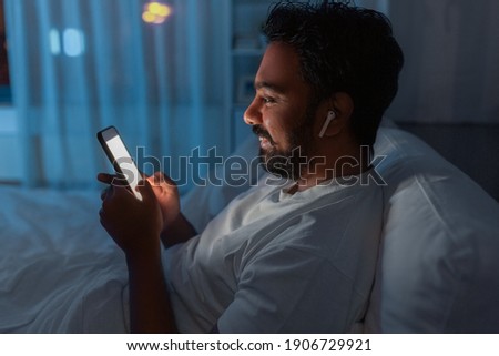 technology, internet, communication and people concept - happy smiling young indian man with smartphone and earphones listening to music in bed at night