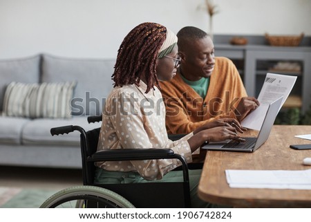 Side view portrait of young African-American woman using wheelchair working from home with husband helping her copy space Royalty-Free Stock Photo #1906742824