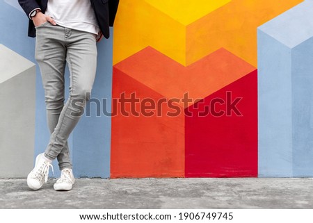 Crop anonymous male in trendy gray skinny jeans and white sneakers holding hand in pocket while leaning against colorful wall Royalty-Free Stock Photo #1906749745