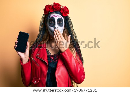 Woman wearing day of the dead costume holding smartphone showing screen covering mouth with hand, shocked and afraid for mistake. surprised expression 