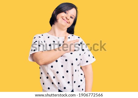 Brunette woman with down syndrome wearing casual clothes cheerful with a smile of face pointing with hand and finger up to the side with happy and natural expression on face 