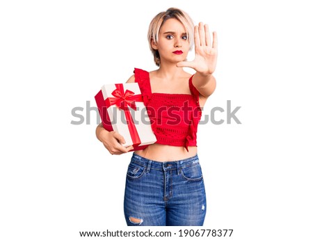 Young beautiful blonde woman holding gift with open hand doing stop sign with serious and confident expression, defense gesture 