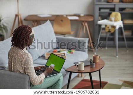 Back view portrait of young African-American woman using laptop with green screen and holding credit card while sitting on sofa at home, copy space