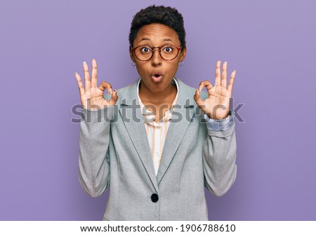 Young african american woman wearing business clothes looking surprised and shocked doing ok approval symbol with fingers. crazy expression 