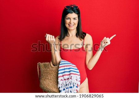 Young hispanic woman wearing swimsuit holding summer wicker handbag smiling happy pointing with hand and finger to the side 