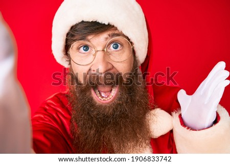 Handsome young red head man with long beard wearing santa claus costume taking a selfie celebrating victory with happy smile and winner expression with raised hands 