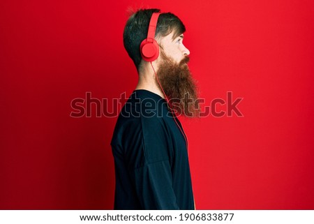 Redhead man with long beard listening to music using headphones looking to side, relax profile pose with natural face with confident smile. 