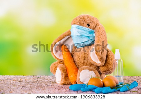 Rabbit on green background with mask for coronavirus, eggs, big carrot, protective gloves and disinfectant around the composition. Happy easter and stay home.