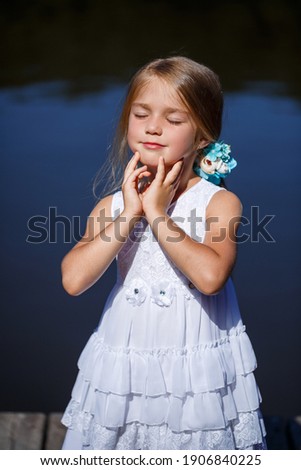 A cute little girl 6 or 7 years old with long blond hair, she is dressed in a ballet summer dress, she is standing on the background of the river