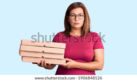 Middle age latin woman holding delivery pizza box thinking attitude and sober expression looking self confident 