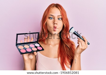 Young redhead woman holding makeup and brush making fish face with mouth and squinting eyes, crazy and comical. 
