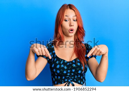 Young beautiful redhead woman wearing casual clothes over blue background pointing down with fingers showing advertisement, surprised face and open mouth 