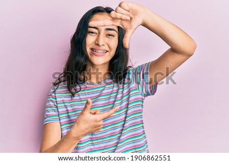 Hispanic teenager girl with dental braces wearing casual clothes smiling making frame with hands and fingers with happy face. creativity and photography concept. 