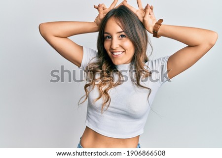 Young brunette woman wearing casual white t shirt posing funny and crazy with fingers on head as bunny ears, smiling cheerful 