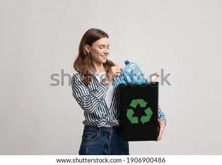 Waste Recycling Concept. Smiling Young Woman Holding Black Container With Plastic Bottles And Green Recycle Sign, Millennial Eco-Friendly Lady Standing Over Gray Background In Studio, Copy Space