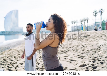 Hispanic female fitness instructor drinking water for refreshing during workout training with blurred male colleague, determined woman in tracksuit holding bottle getting ready for cardio exercising