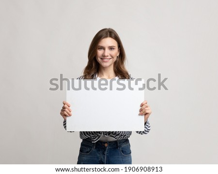 Beautiful Young Woman Holding Blank Placard With Copy Space For Advertisement. Smiling millennial lady demonstrating white empty billboard with place for text, standing over grey studio background Royalty-Free Stock Photo #1906908913